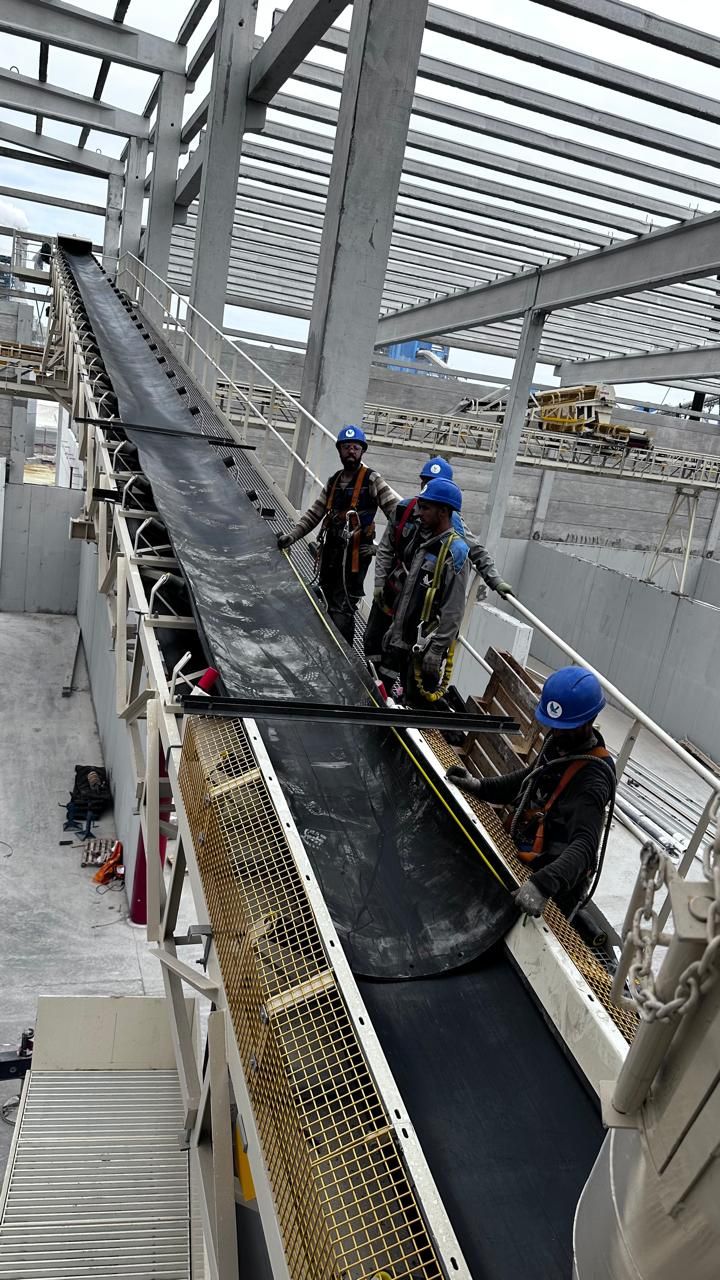 Trois techniciens en casque bleu et harnais tirant une bande neuve le long d'une passerelle de convoyeur métallique, structure industrielle à toit ajouré