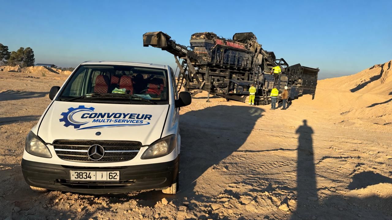 Véhicule utilitaire floqué Convoyeur Multi-Services garé devant un engin mobile de concassage-criblage, techniciens en gilet haute visibilité à l'arrière-plan, site en plein air